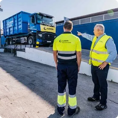 Foto de dos operarios, uno de ellos señalando con su mano hacia un camión de reciclaje.